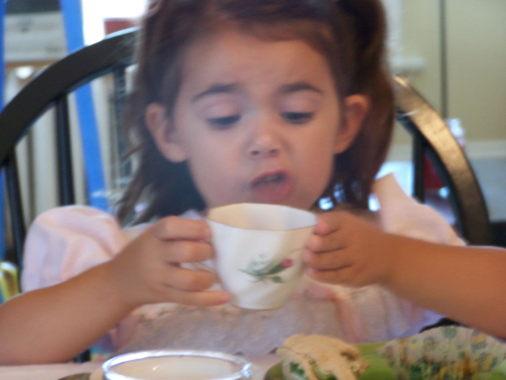 A small girl in a white dress drinking from a china teacup at a table set for a tea party