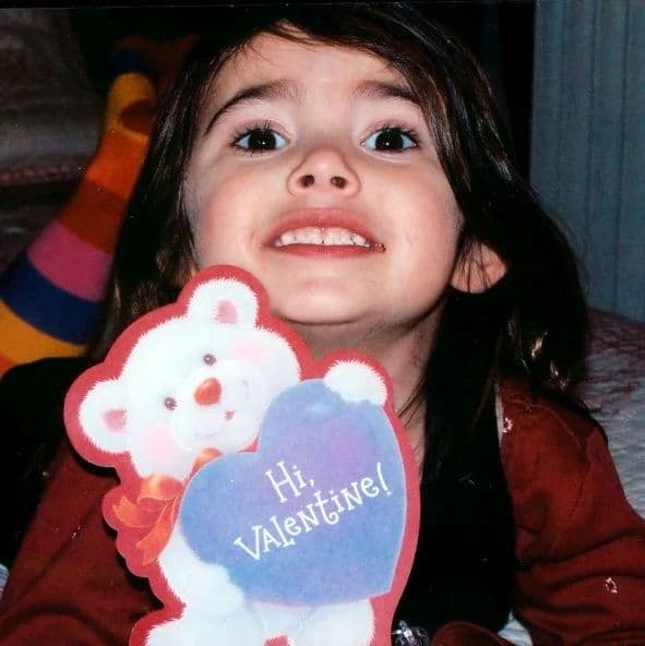 A dark-haired toddler in a red coat holding up a Valentine card with a teddy bear on it