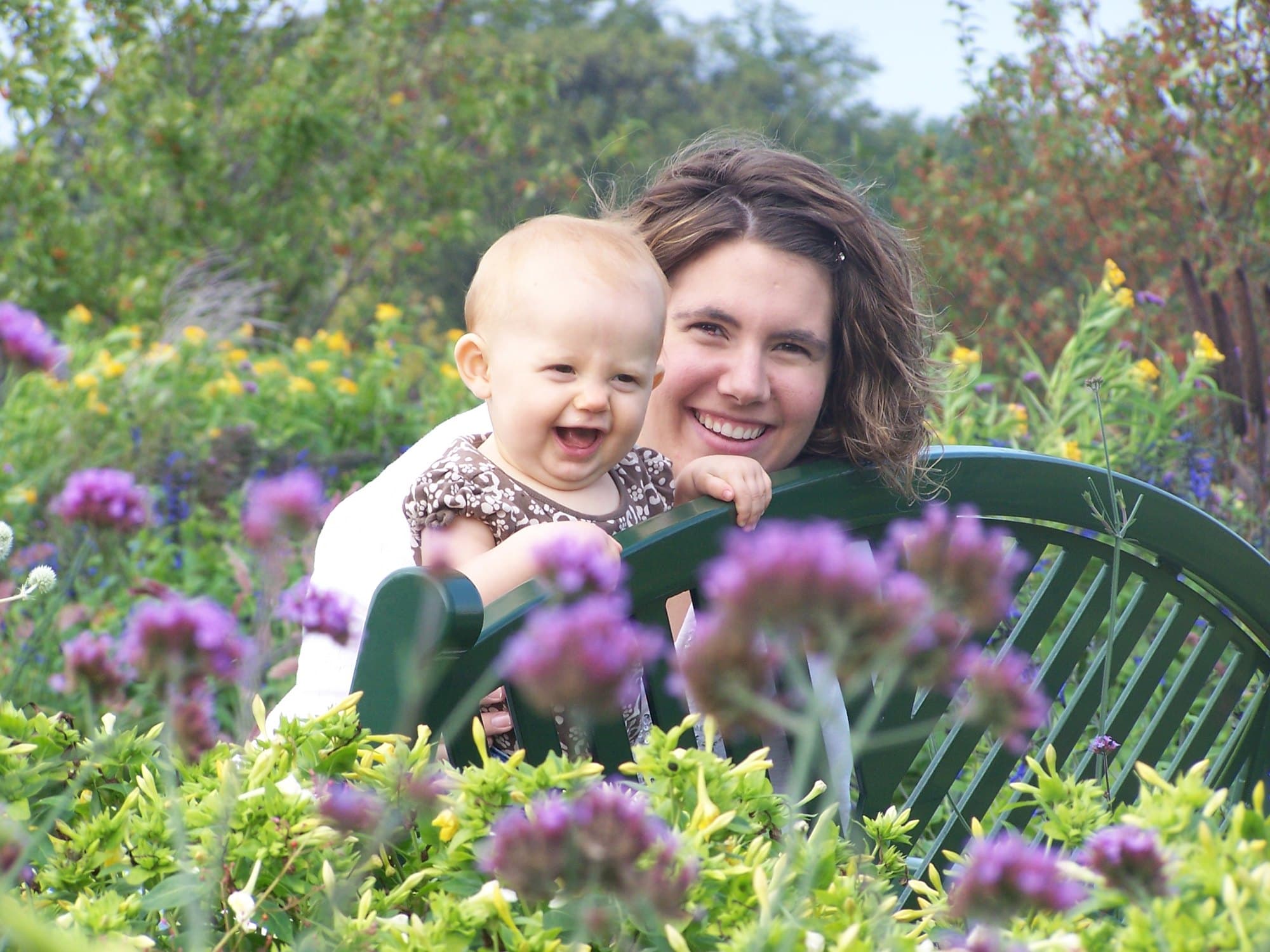 Gretchen and a laughing baby leaning on a garden bench among purple flowers