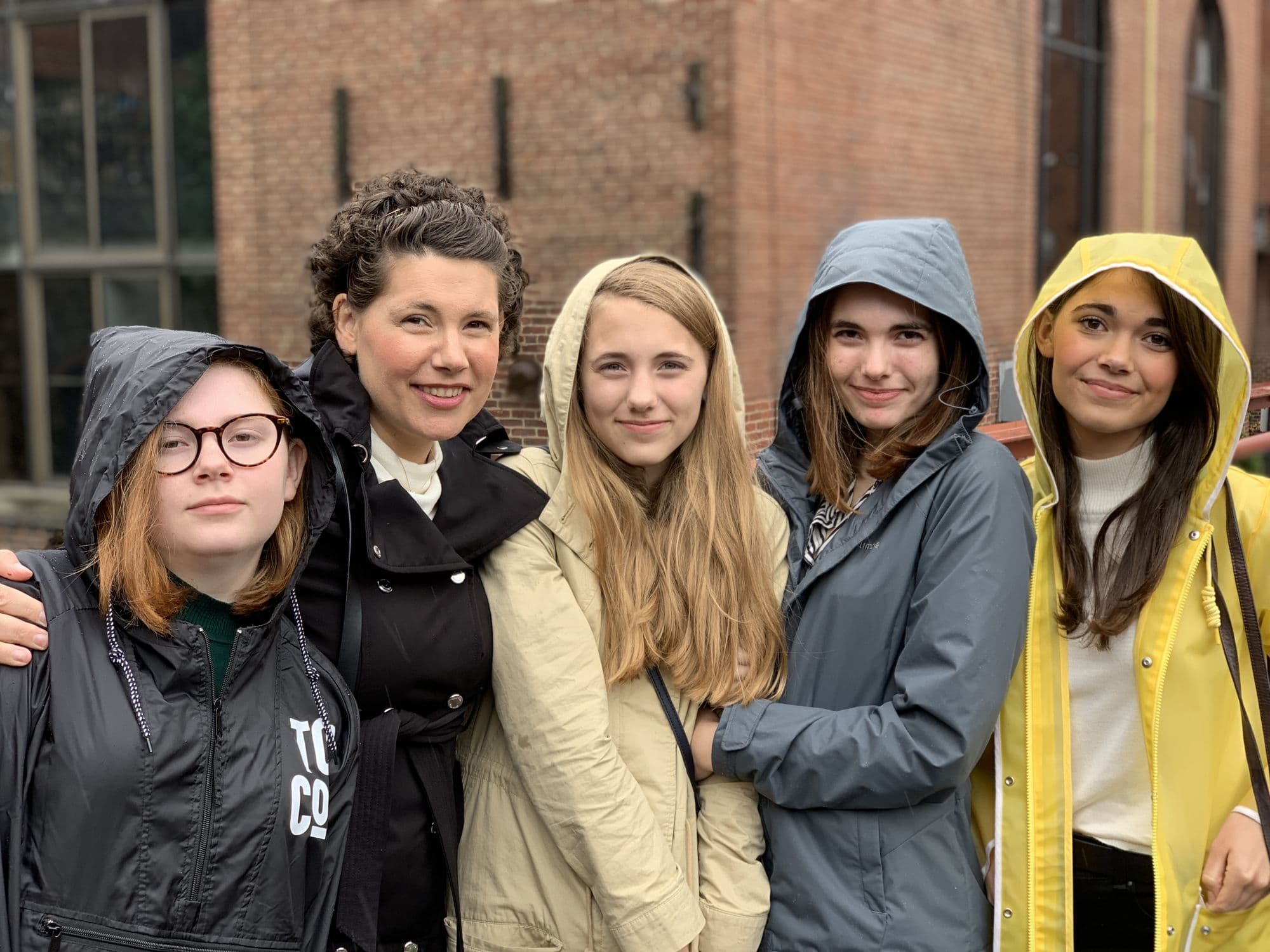 Five people in raincoats pose outside a brick building