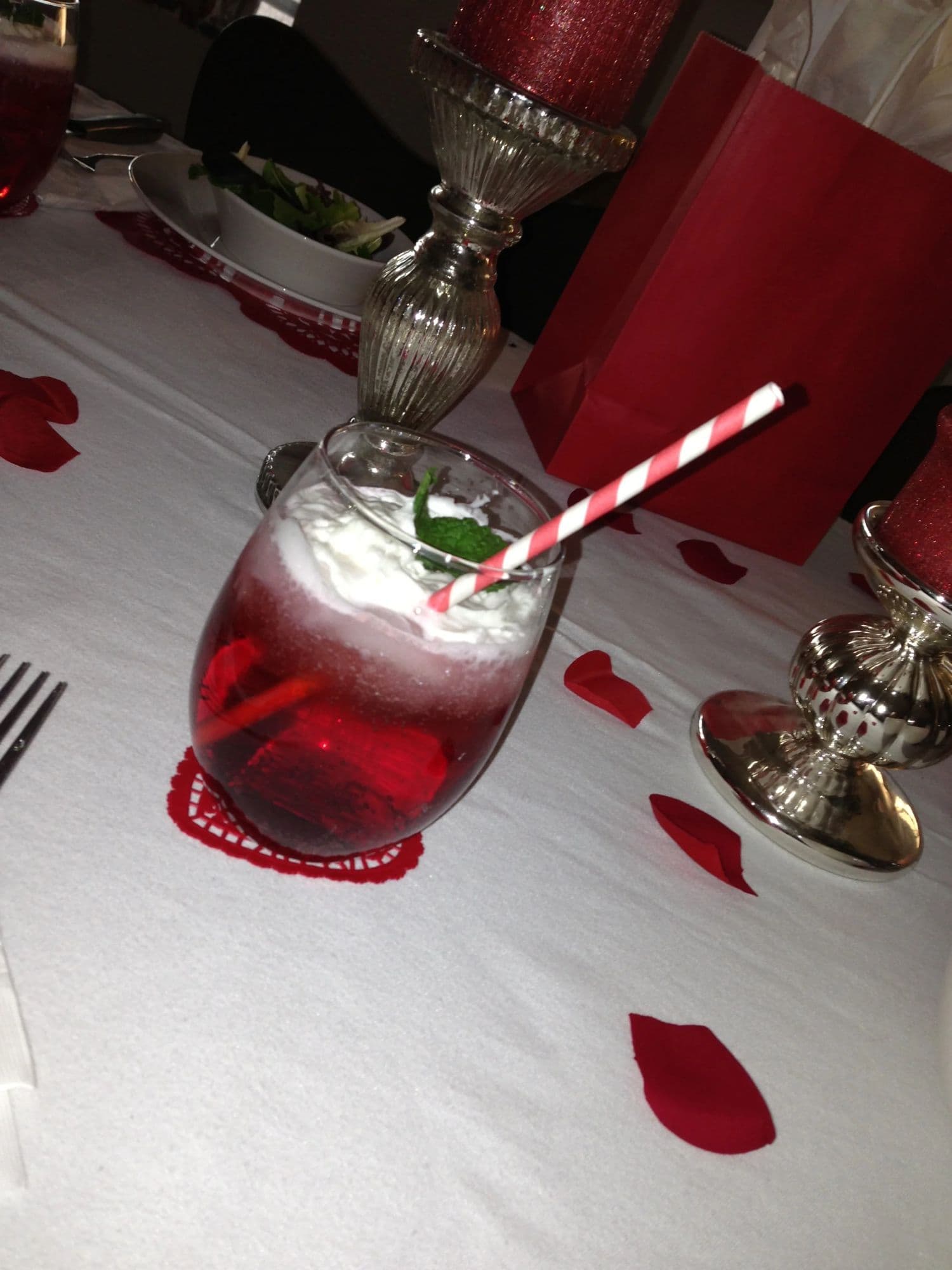 A red drink in a footed glass with a striped straw, rose petals scattered on the tablecloth around it
