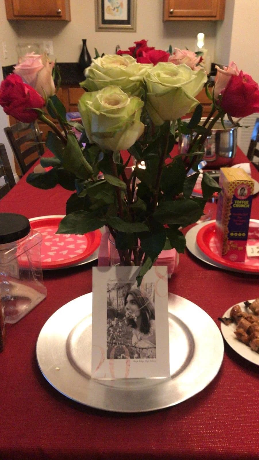 A vase of roses on a set dinner table, a framed photograph propped against a charger plate