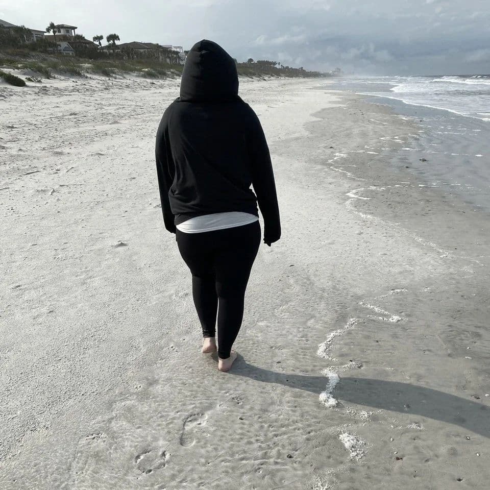 A person in a black hooded sweatshirt walking away along an empty beach under a grey sky