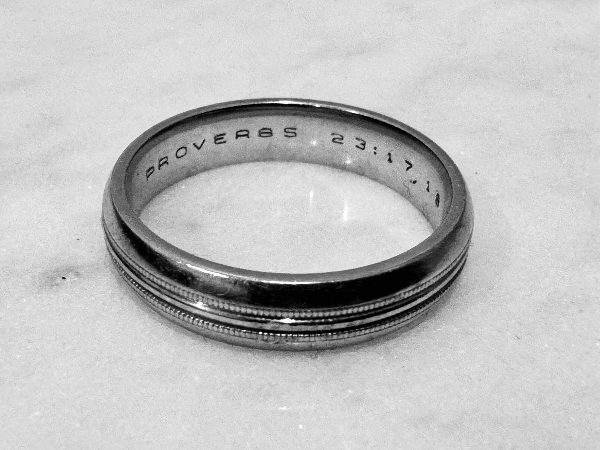 A wedding band photographed on a marble surface, Proverbs 23:17 engraved inside it